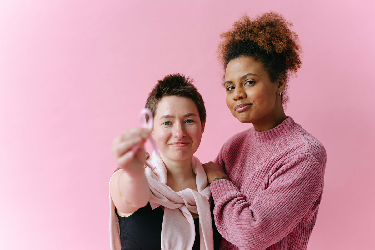 Two women standing together; one holds a pink breast cancer awareness ribbon forward, pink background.